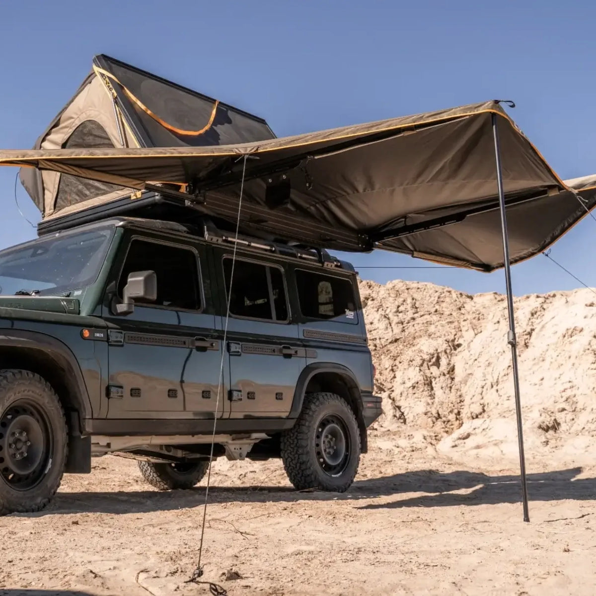 A dark green off-road vehicle equipped with a rooftop tent and the CVT 180 Degree Awning, which offers instant shade, is parked on sandy terrain beneath a clear blue sky.
