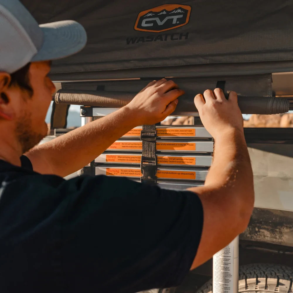 A person adjusts the ladder of a CVT Wasatch Hardshell Tent mounted on a vehicle, preparing for an overland adventure, with the distinctive CVT branding visible on the tent fabric.