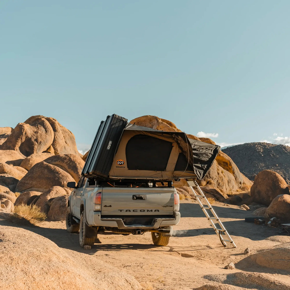 A pickup truck equipped with a CVT Wasatch Hardshell Tent is parked among large desert rocks. A ladder extends from the tent to the ground under a clear blue sky, ready for the next overland adventure.