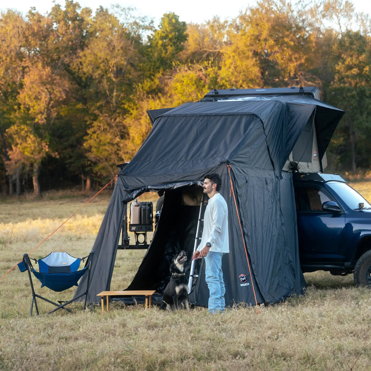 A man stands outside a rooftop tent attached to a blue vehicle in a grassy field with a dog. Camping gear and a chair are nearby. Trees with autumn leaves are in the background.