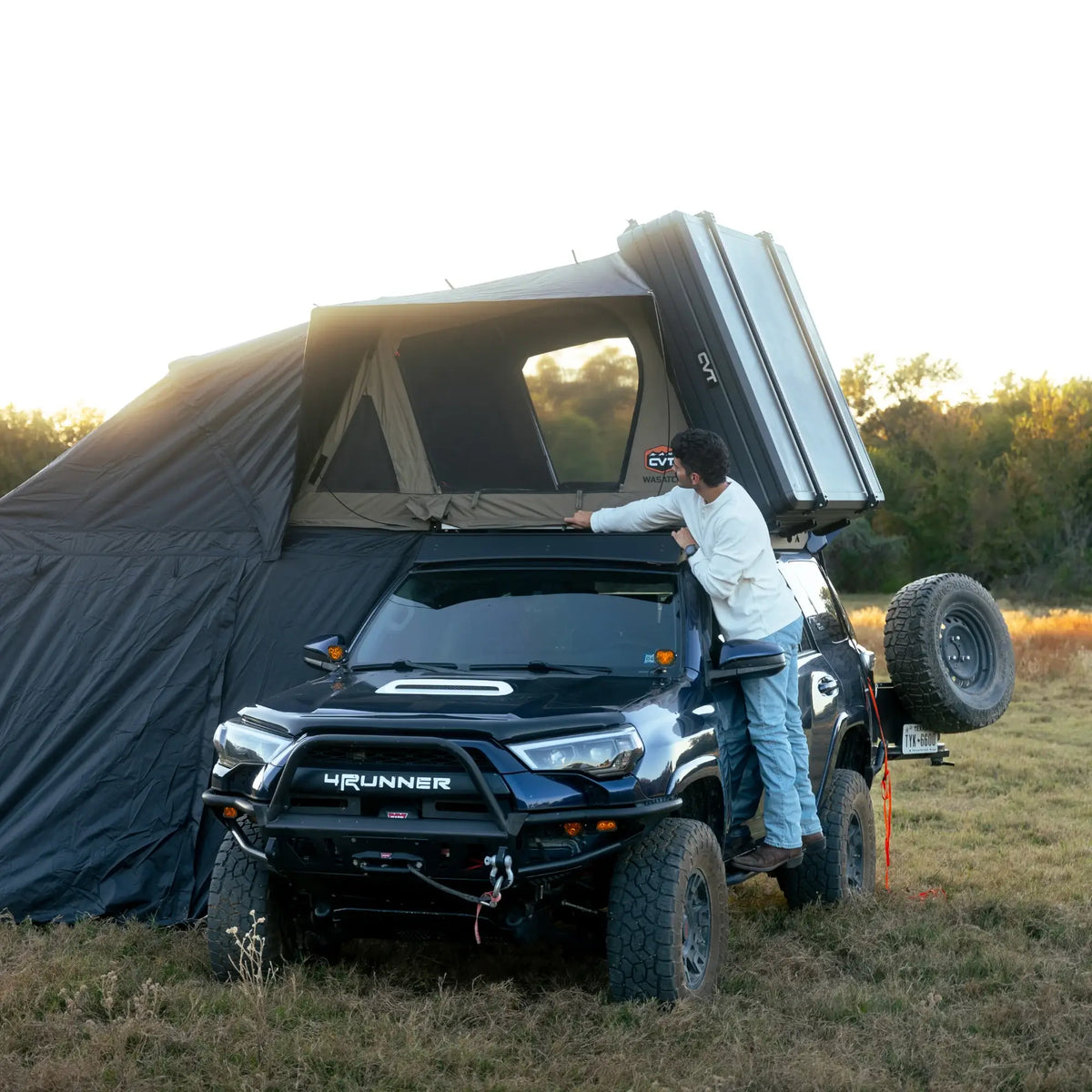 A person stands beside a black SUV installing a CVT Wasatch Annex rooftop tent from CVT, which is attached to the vehicle, in an open grassy field with trees in the background.