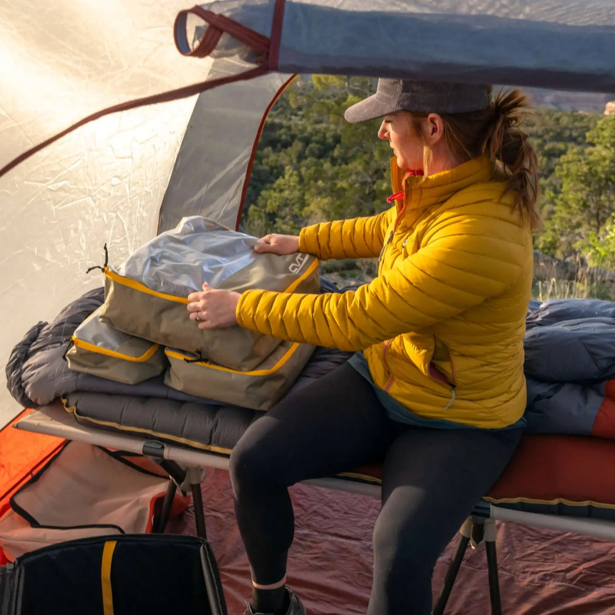 A person in a yellow jacket sits on a cot inside a tent, practicing gear organization with CVT Camping Cubes™ as they arrange bags on top of a water-resistant sleeping bag. Sunlight filters through the tent, and trees are visible outside.