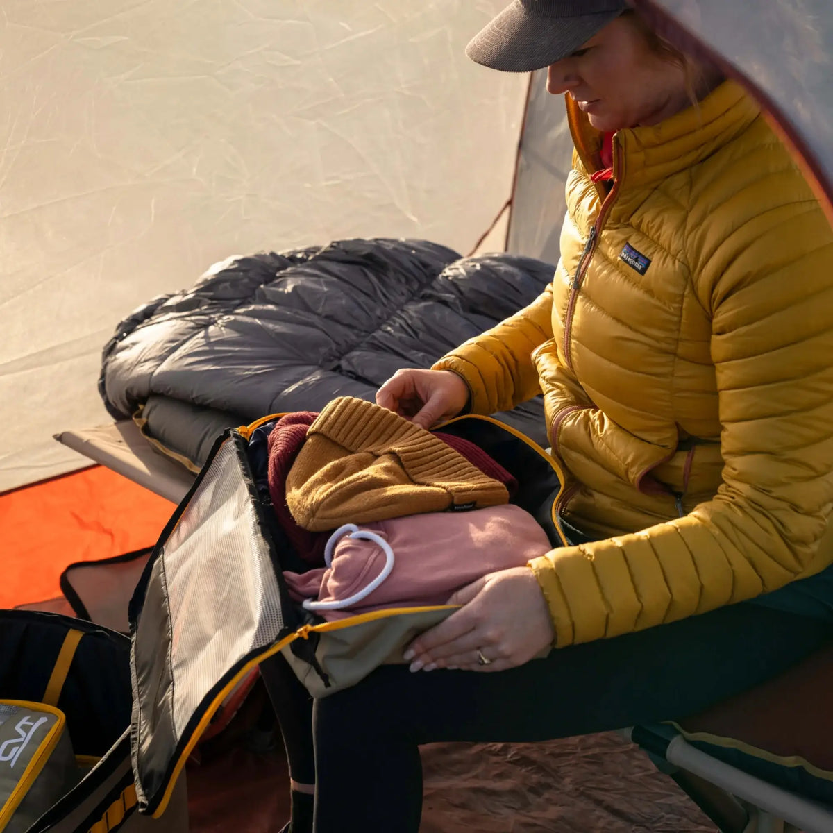 A person in a yellow jacket sits in a tent, skillfully organizing clothes into a CVT Camping Cubes™ for efficient gear organization, with a sleeping bag and other water-resistant camping gear visible in the background.