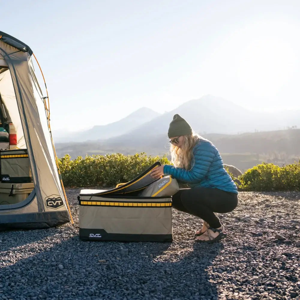 Wearing a blue jacket and beanie, someone opens a CVT Camping Cubes™ water-resistant portable storage box next to a tent, with mountains and lush greenery behind them and sunlight streaming in—ideal for efficient gear organization on every adventure.