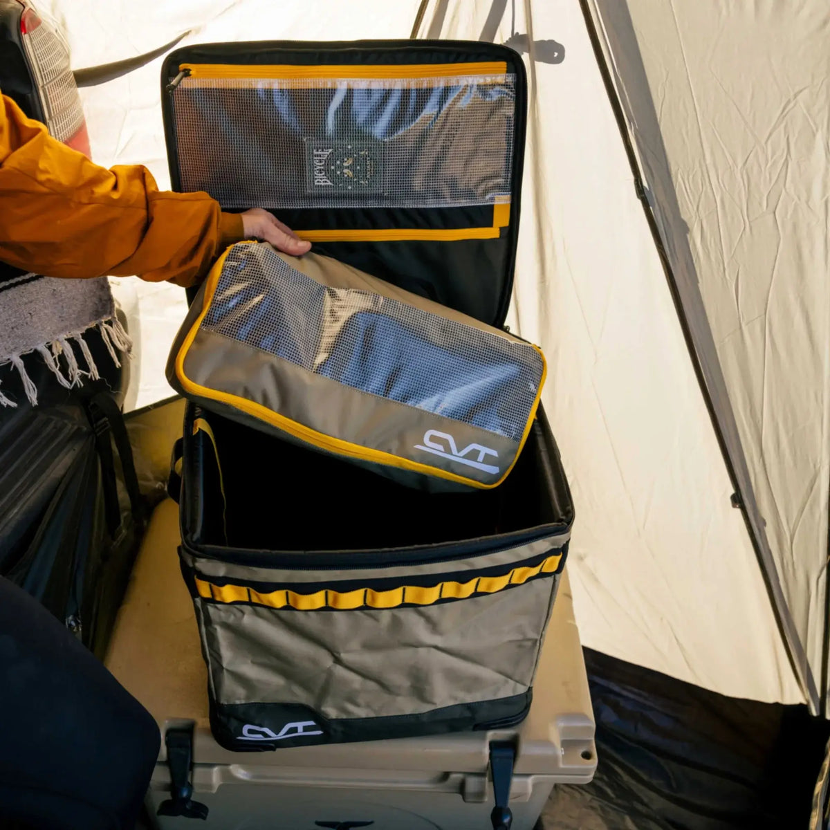 Inside a tent, someone organizes their gear by placing a mesh travel pouch into a square storage bin with yellow trim—both featuring the CVT logo. These Camping Cubes™ by CVT are perfect for keeping your essentials tidy on any camping adventure.