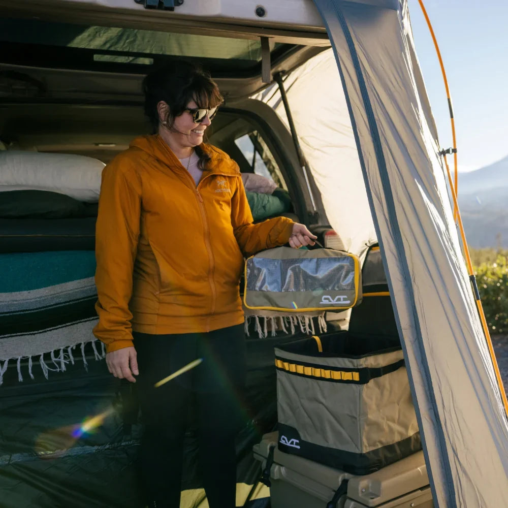A woman in a yellow jacket stands by an open car trunk, holding a CVT Camping Cubes™ packing cube while camping. A tent, bedding, and storage containers from CVT ensure easy gear organization around her in the sunlight.