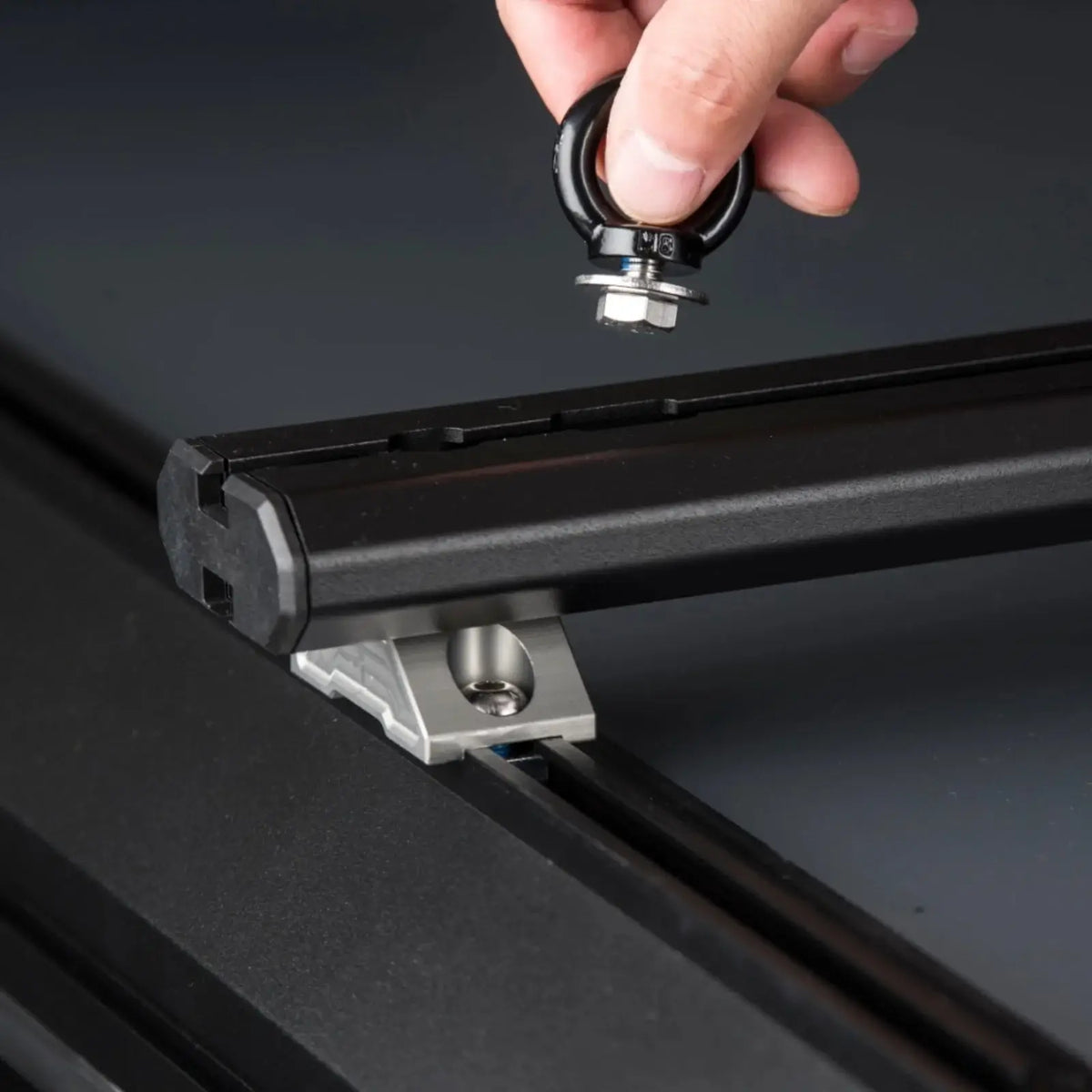 A hand holds a black ring bolt above a black metal rail, preparing to insert it into the aluminum slot of a CVT Mt. Hood/La Sal Cross Bar - 2 Pack, which features a slotted bracket and visible hardware—perfect for securing hardshell tents and other mounting accessories.