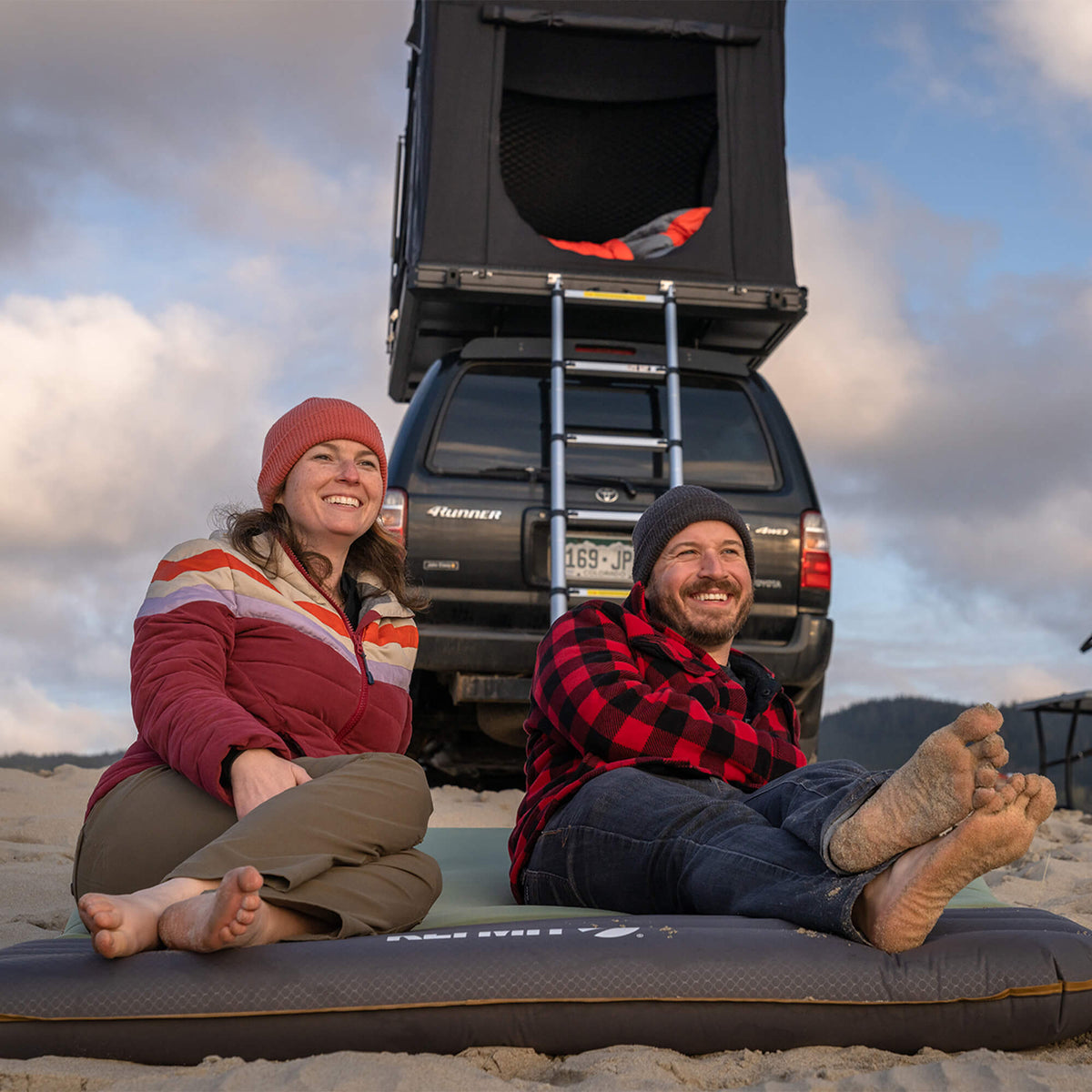 Two people sit barefoot on a mat at the beach, smiling. Behind them is a black SUV with a rooftop tent and ladder, ready for adventure. Their Klymit Klymaloft™ Double Sleeping Pad provides extra comfort as they enjoy the view under a partly cloudy sky.