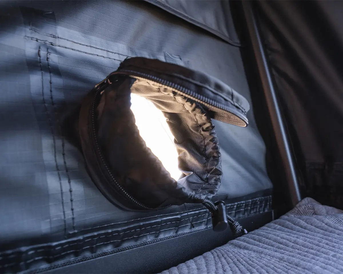 A round zippered window in a dark fabric tent is partially open, letting bright light shine through. The window has a fabric flap and is next to a gray padded surface.