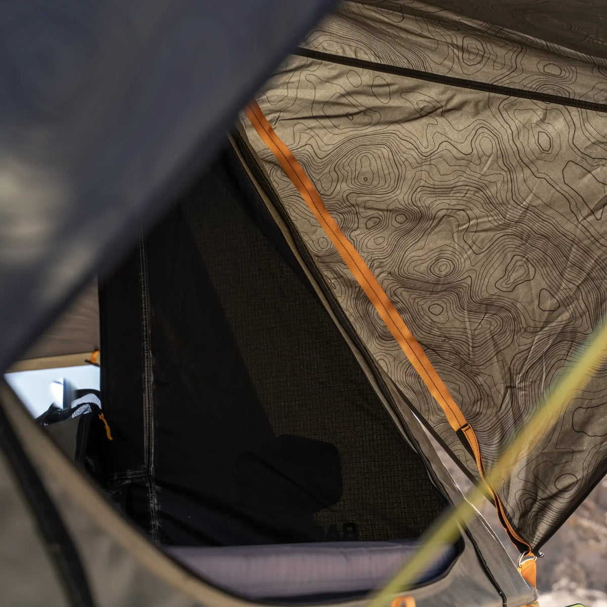 Close-up of the CVT La Sal Hardshell Tent showcasing its side-open design, partially unzipped to reveal a sleeping pad inside. The tent fabric displays a topographic map pattern, while orange zippers accentuate the opening—highlighting the spacious comfort and innovative rooftop experience of this tent.