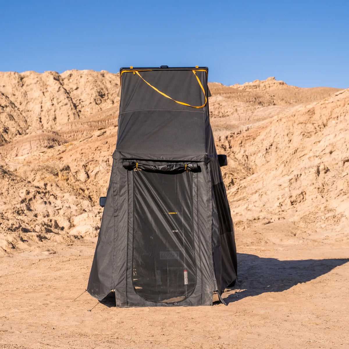 The CVT Mt. Hood Annex, a tall black pop-up privacy tent, stands on sandy ground with tan rocky hills and a clear blue sky in the background—serving perfectly as an outdoor gear storage solution or as a versatile tent annex for added convenience.