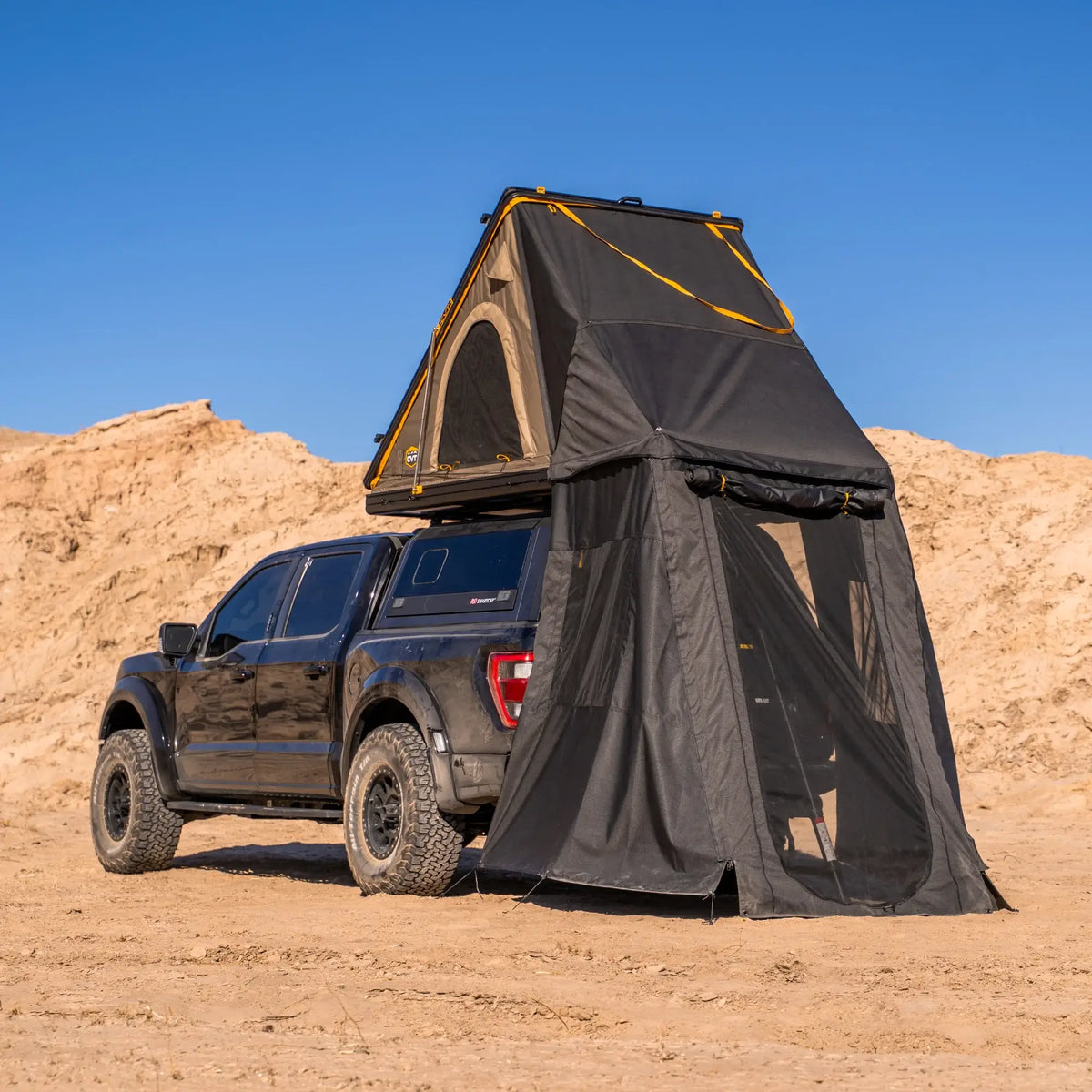 A black pickup truck is parked on sand under a clear blue sky, with a roof tent extended upward and a spacious CVT Mt. Hood Annex attached, providing ample outdoor gear storage.