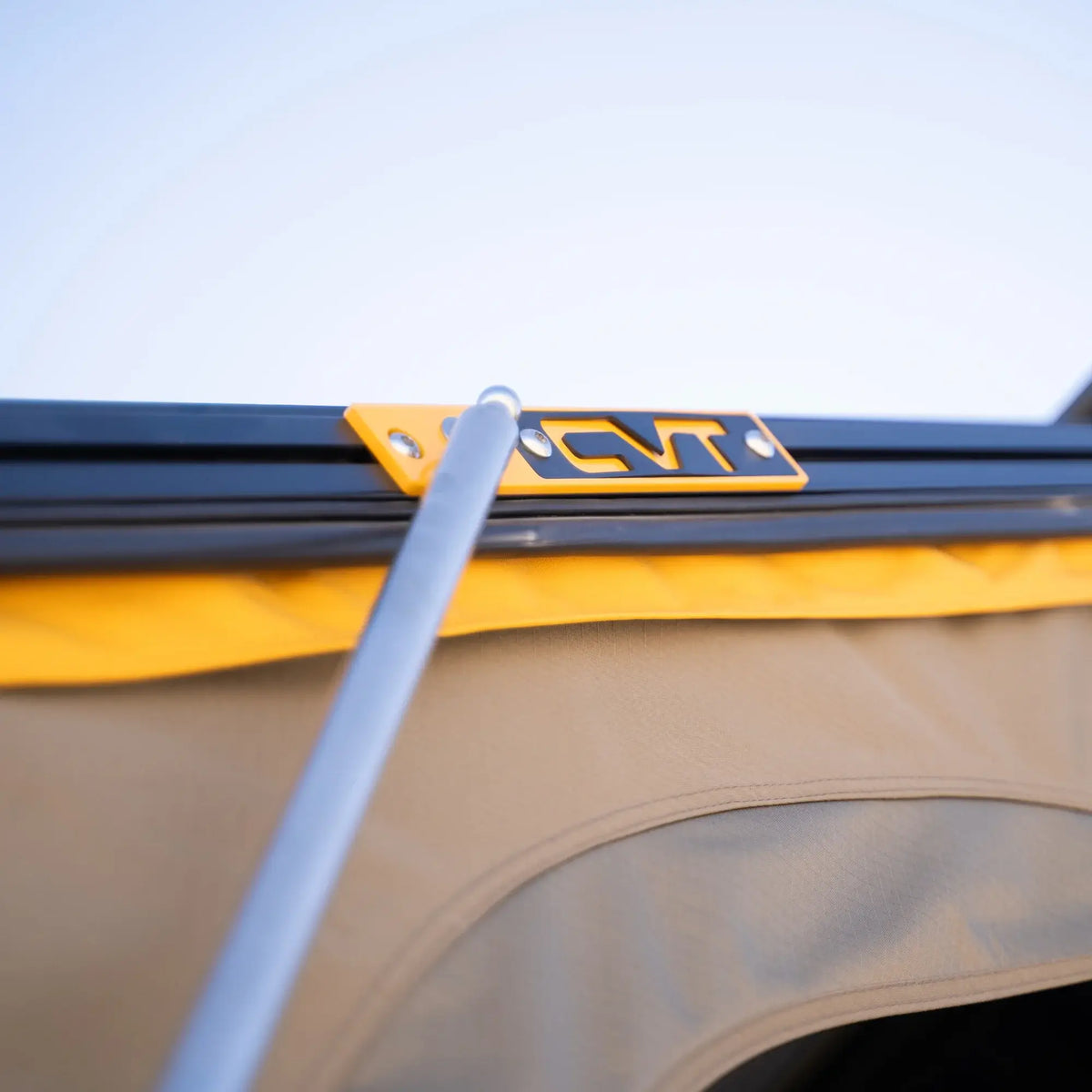 A close-up of a tent pole connected to a black metal frame with a yellow CVT bracket, with tan and yellow fabric from the Mt. Hood 2 Hardshell Tent by CVT visible underneath and a clear sky in the background—showcasing the durability and thoughtful design that make this rooftop tent ideal for your outdoor adventures.