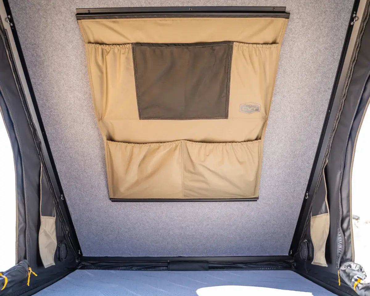 Interior view of a tent roof with beige storage pockets, one with a black mesh section, attached to light gray fabric walls. The photo is taken from inside, looking upward at the storage area.