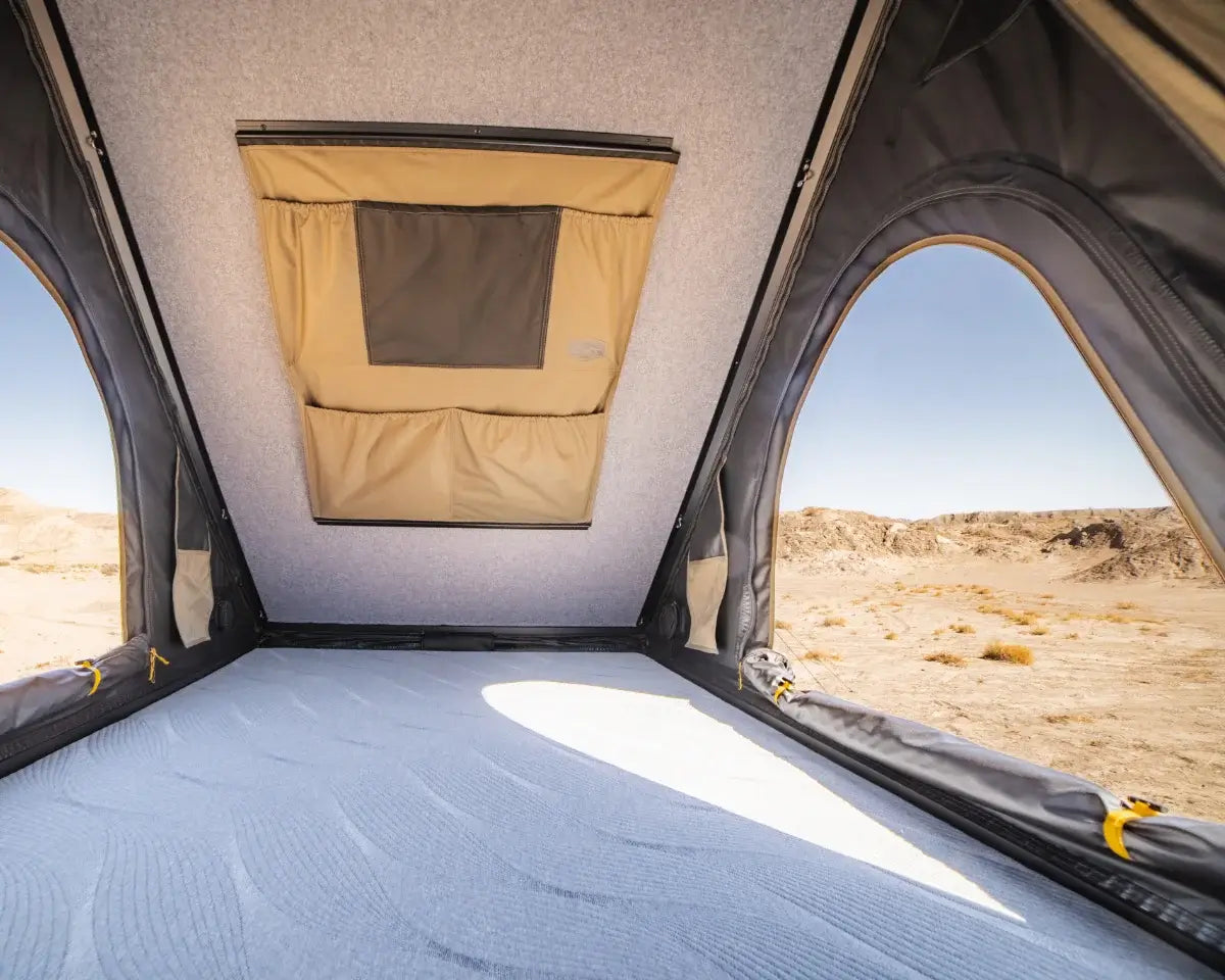 View from inside a rooftop tent, showing a gray mattress, two side windows, and a ceiling vent, with a desert landscape visible outside.