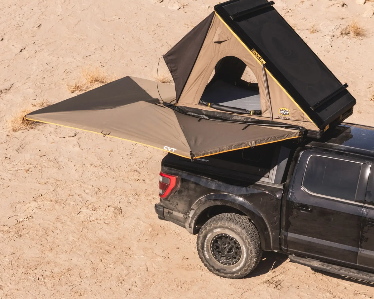 A black pickup truck with a rooftop tent and an extended awning is parked on sandy terrain, ready for camping in a desert environment.