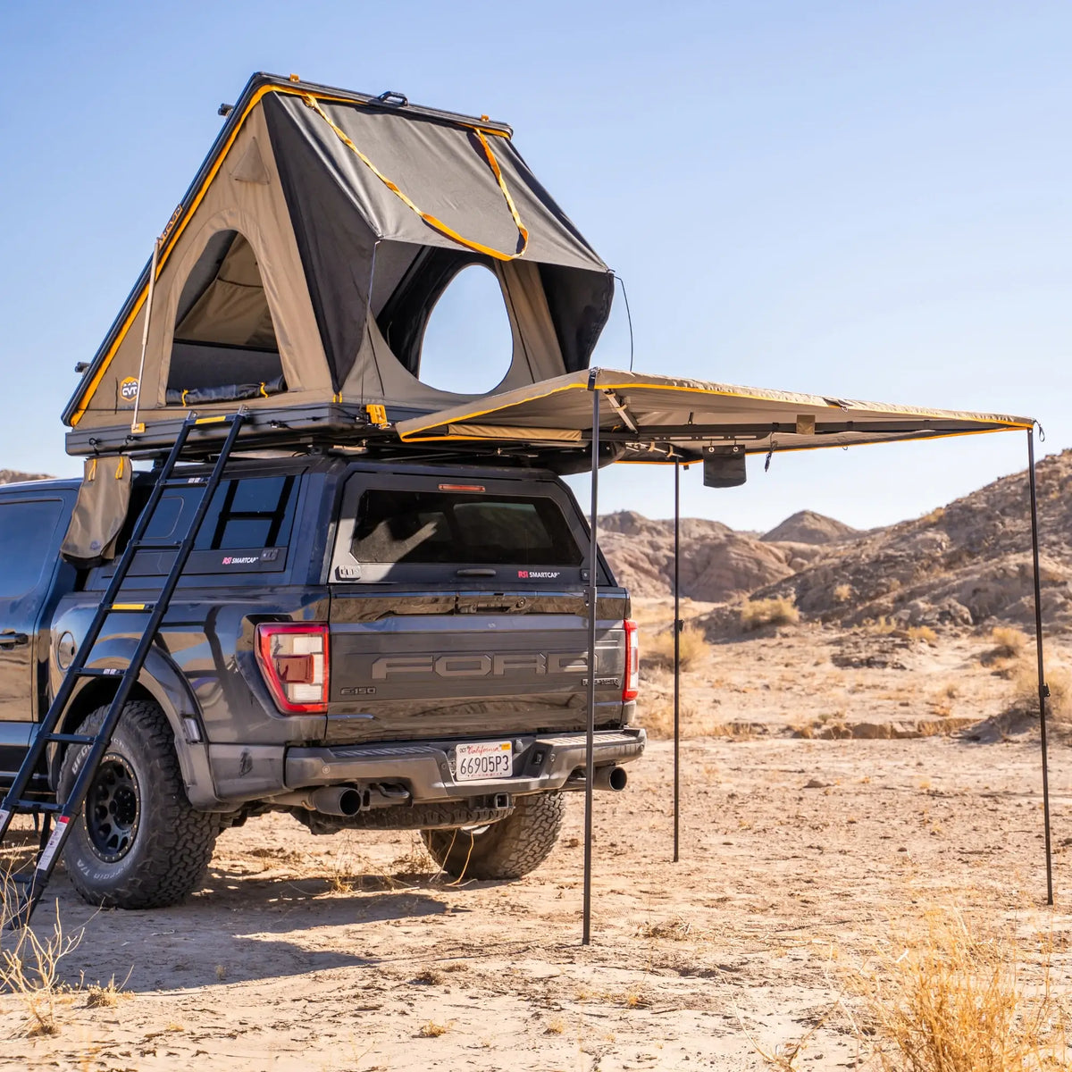 A truck equipped with the CVT Mt. Hood 2 Hardshell Tent and awning is parked on sandy terrain in a desert landscape, with mountains in the background under a clear blue sky—perfect for outdoor adventures.