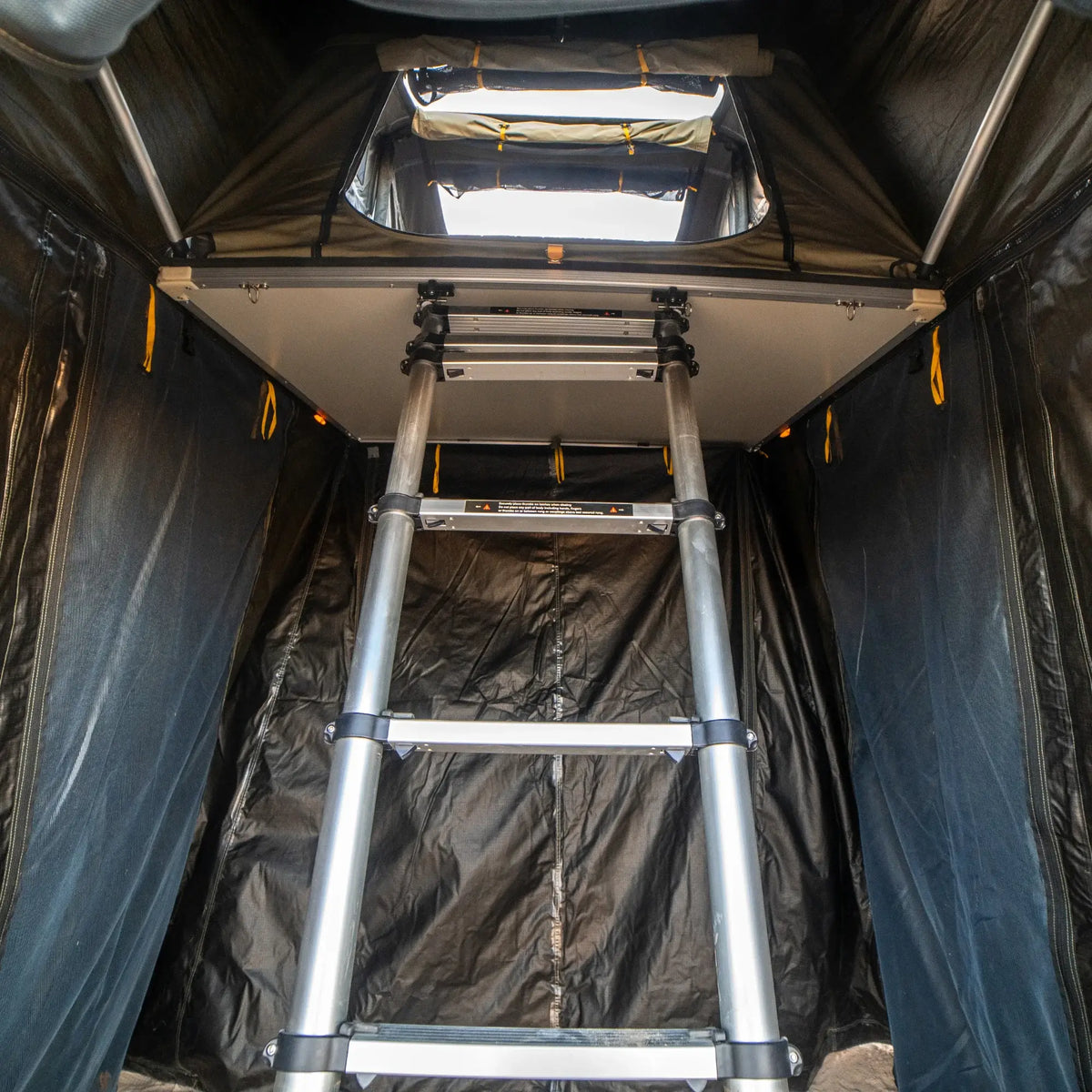 View from inside an enclosed tent space, looking up at a metal ladder leading to the rooftop tent opening, with dark fabric walls on both sides—the ideal setup for attaching your CVT Pioneer Annex.