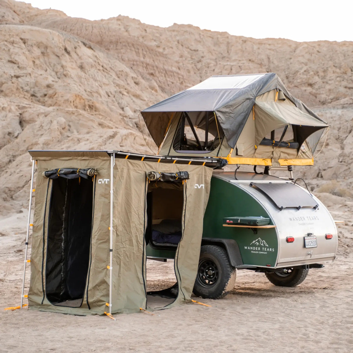 A teardrop camper equipped with CVT Pioneer Awning Walls made from waterproof ripstop material, along with a rooftop tent, is parked in a desert landscape with rocky hills in the background.