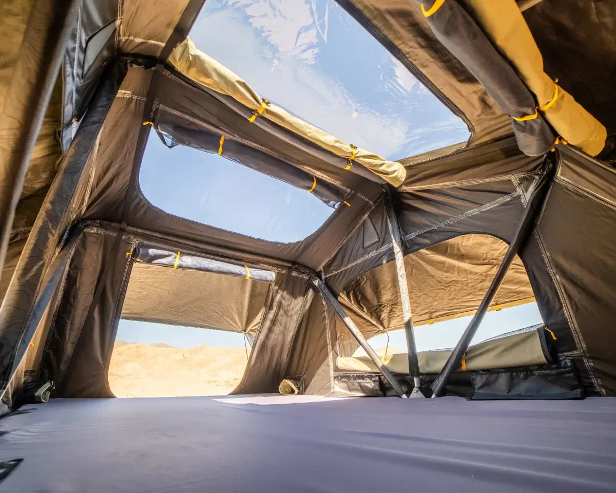 View from inside a rooftop tent, showing windows, sunlight, and clear sky through the ceiling. Mattresses and support poles are visible, with a sandy landscape outside the windows.
