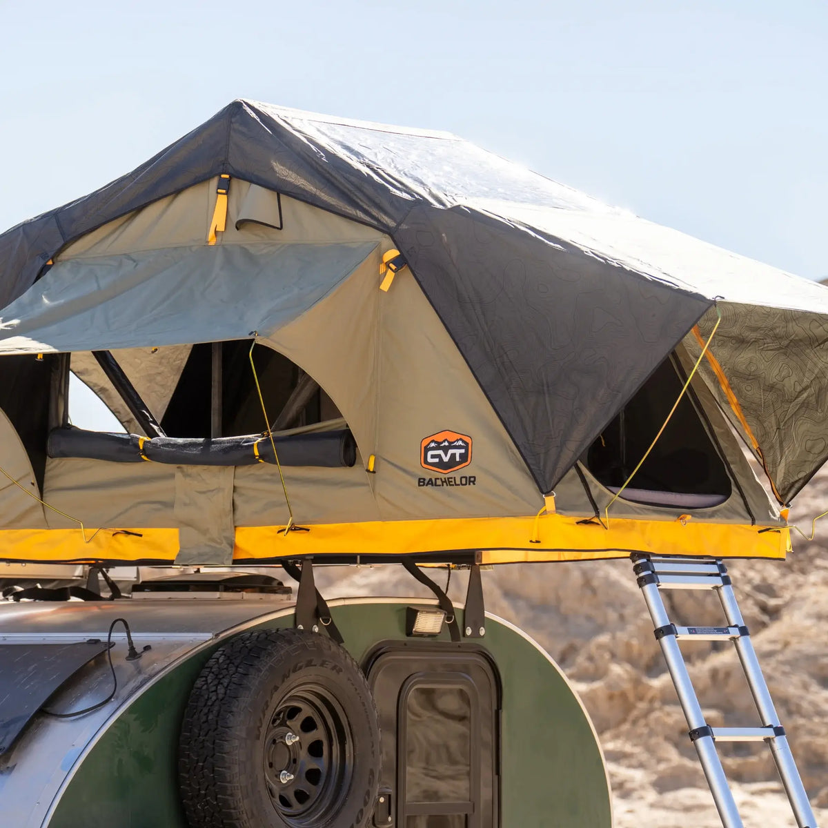 A CVT Pioneer 2 Softshell Tent with a black and yellow cover is set up on top of a green camper trailer amid a rocky, desert landscape—perfect for overland adventures. A folded ladder leads up to the tent’s entrance.