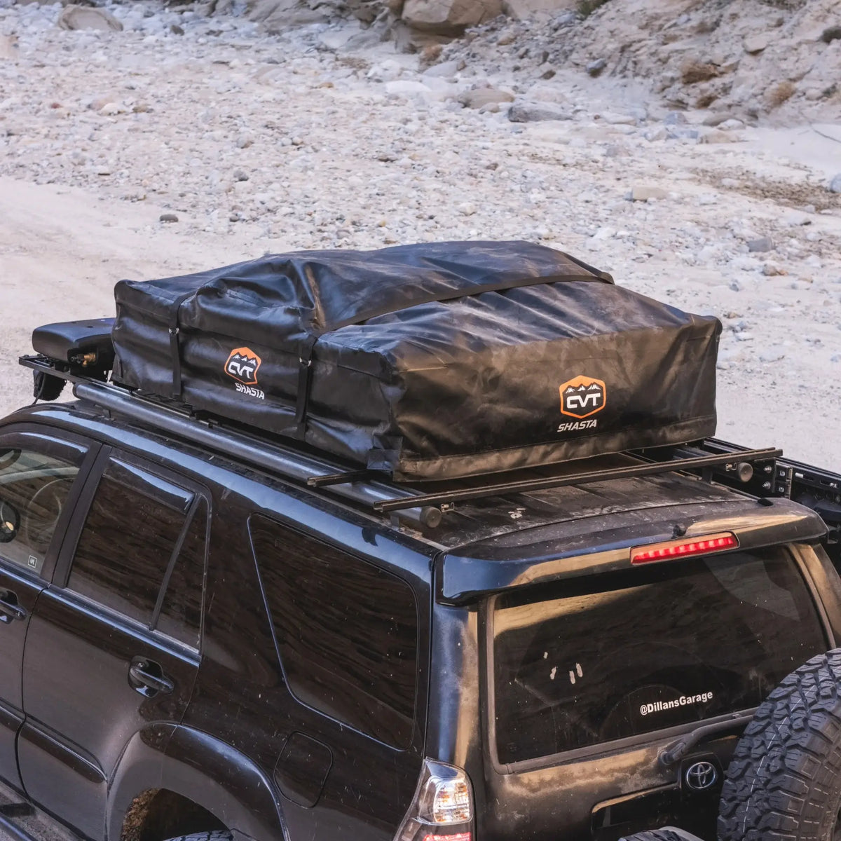 A black SUV with a CVT Pioneer 2 Softshell Tent mounted on a roof rack is parked on rocky, sandy terrain—perfect for overland adventures. The soft-shell tent is folded and secured in a sleek black protective case.