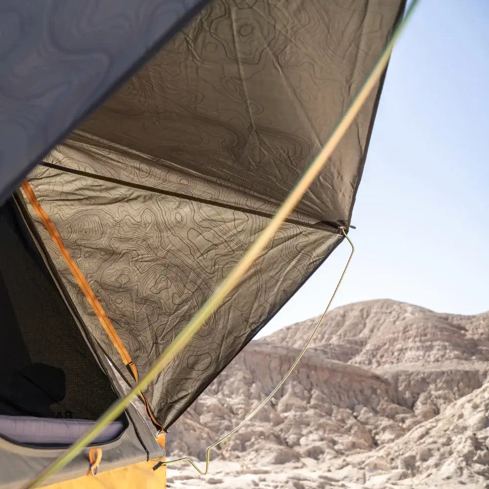 Close-up of a pitched tent featuring CVT’s Pioneer Spring Rod with corrosion-resistant yellow tent poles and topographic pattern fabric, set against rocky desert hills under a clear sky.