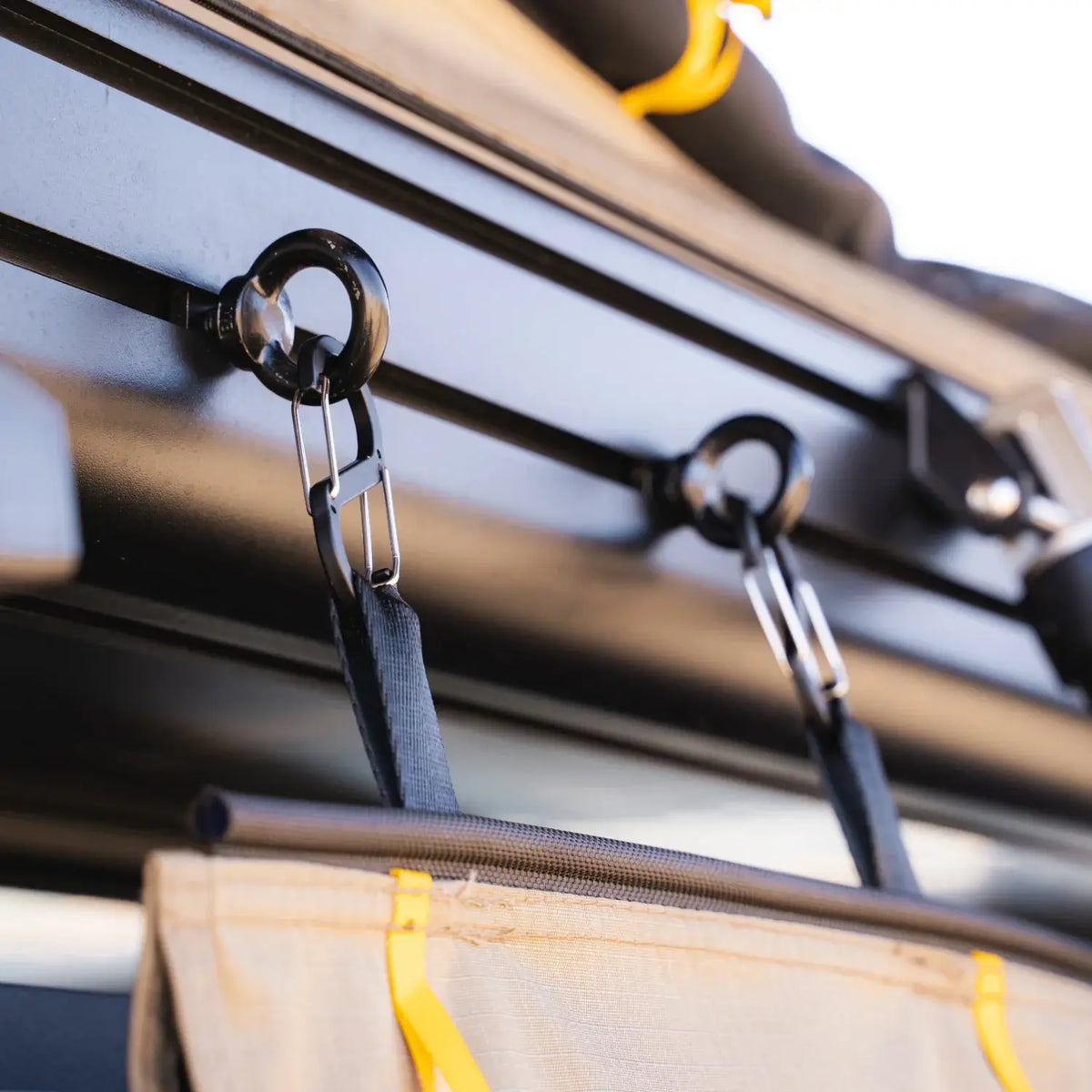 Close-up of two black metal hooks attached to a rail, each supporting a strap connected to a beige fabric item with yellow trim—the CVT Mt. Hood/La Sal Shoe Bag—designed for convenient rooftop tent shoe storage as part of outdoor or camping gear.