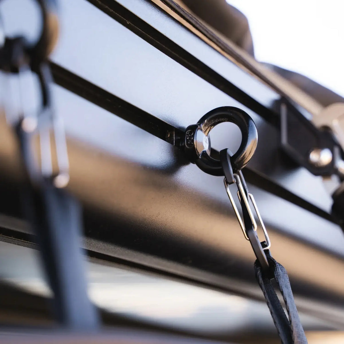 Close-up of a black metal ring and carabiner fastened to the rail of a roof rack with secured straps on a vehicle. Perfect for attaching accessories such as the CVT Mt. Hood/La Sal Shoe Bag or other rooftop tent shoe storage solutions, this setup keeps your essentials organized while traveling. The background is softly blurred.