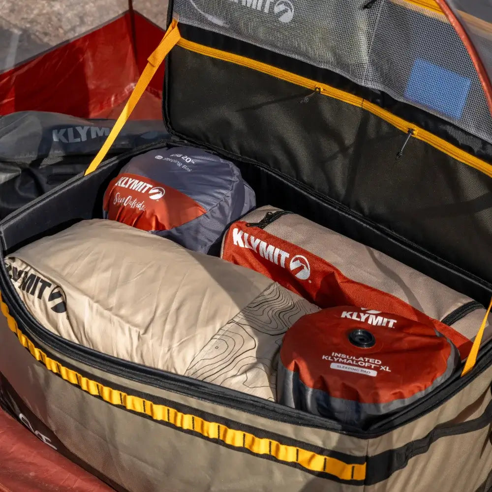 The CVT Soft Side Storage™ bag, packed with multiple Klymit sleeping bags and gear arranged neatly inside, is placed outdoors with a glimpse of a red tent in the background—providing convenient, water-resistant storage for all your adventure essentials.