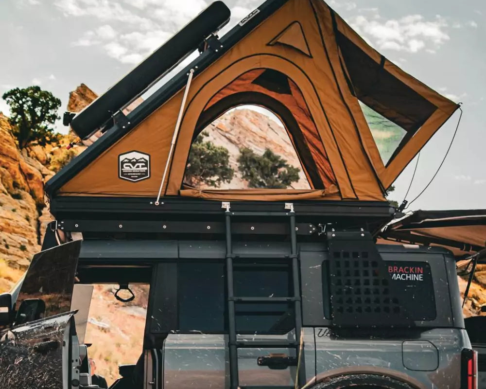 A rugged off-road vehicle with a rooftop tent is parked in a rocky, desert landscape. The tent is open, with a ladder leading up, and the vehicle doors are open, suggesting an outdoor camping adventure.