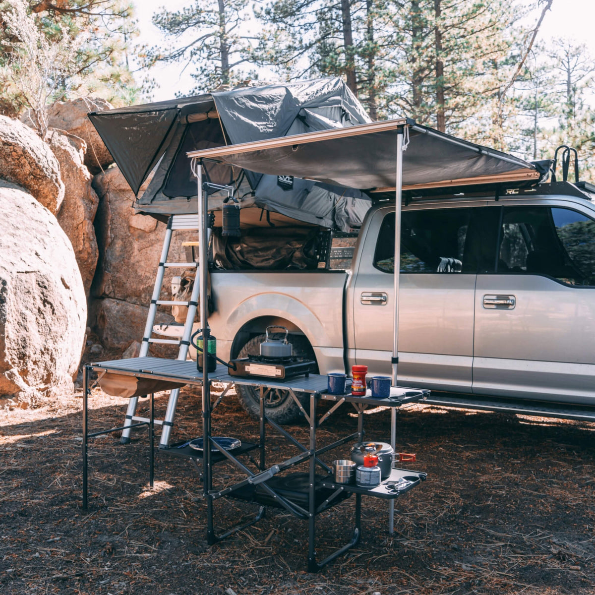 A silver truck equipped with a rooftop tent and the CVT Pioneer Awning - 79″ Medium is parked at a campsite. Beside the truck, a folding table arranged with cooking gear, cups, and food items sits under tall trees near large rocks, providing a comfortable outdoor shelter for relaxing and dining.