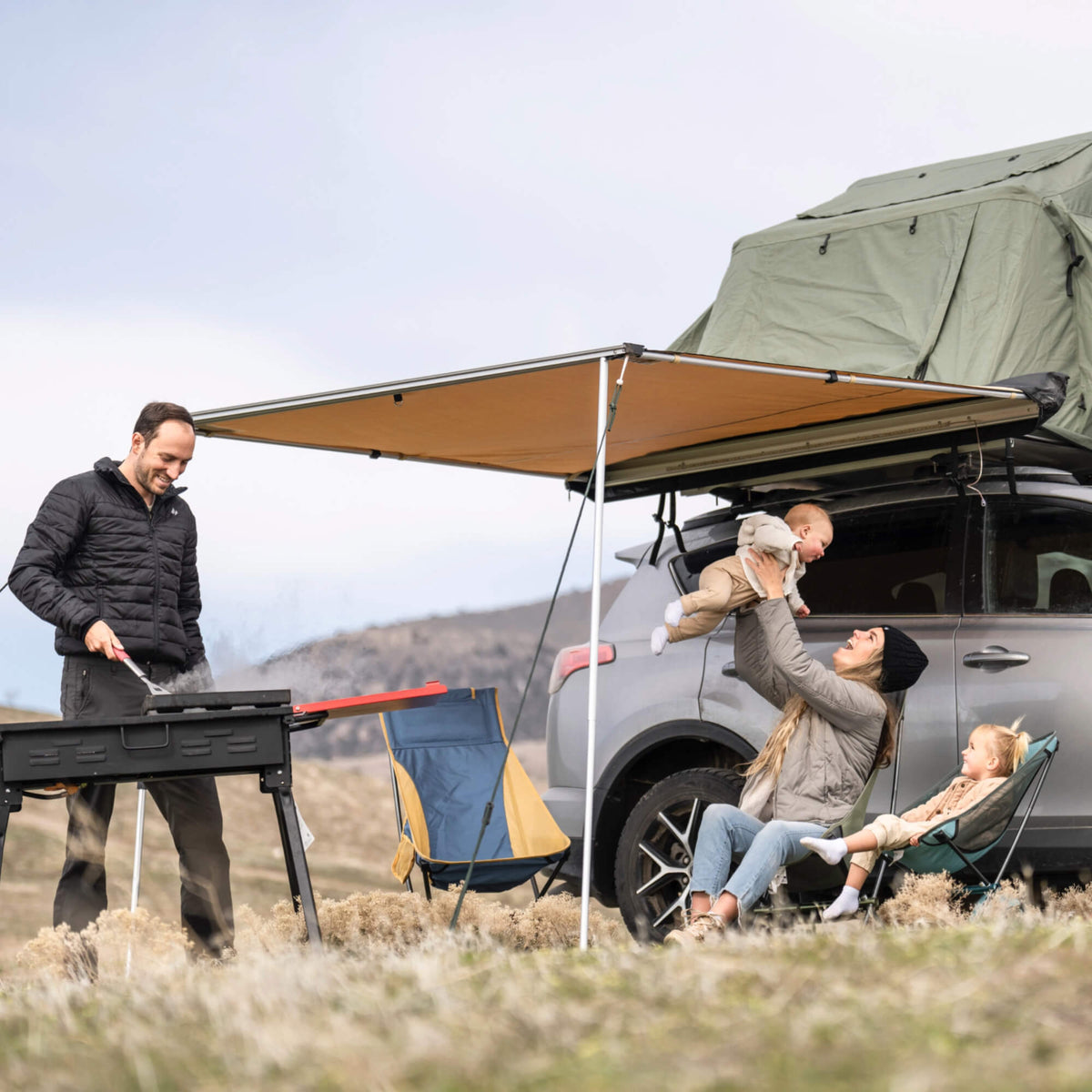 A man grills food by a car equipped with a rooftop tent and the CVT Pioneer Awning - 79″ Medium, while a woman sitting in a chair lifts a baby with a young child nearby. They are outdoors in a grassy, mountainous area, enjoying the added comfort of their CVT Pioneer Awning shelter.