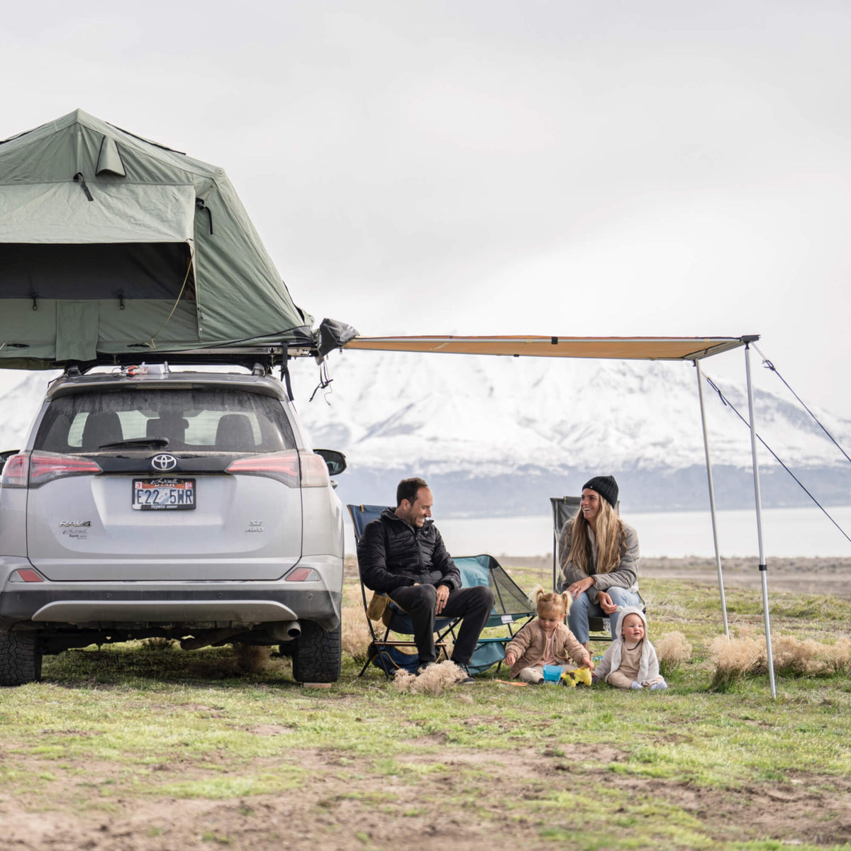 A family with two young children and a dog sits beside their car, which is equipped with a rooftop tent and a CVT Pioneer Awning - 79″ Medium, as they camp by a lake with snow-capped mountains in the background on a cloudy day.