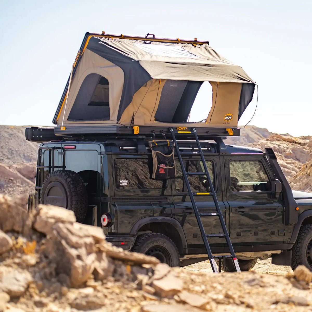 A black off-road vehicle is parked on rocky desert terrain under a clear sky, equipped with the CVT La Sal Hardshell Tent featuring a side-open design and ladder, providing spacious comfort for adventurous travelers.