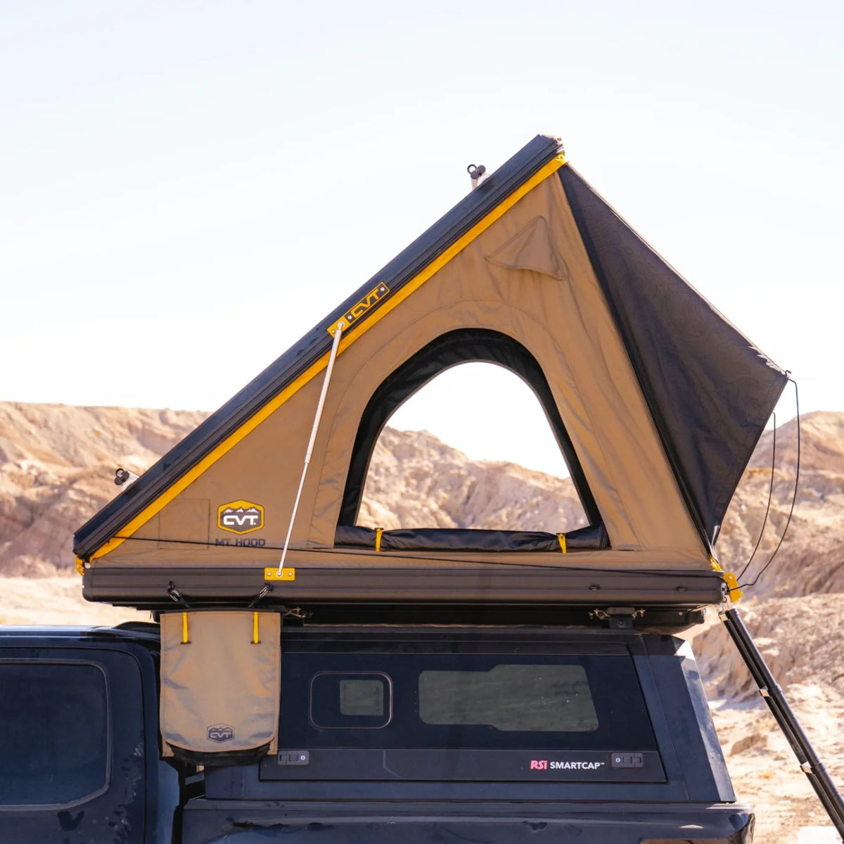 A CVT Mt. Hood 2 Hardshell Tent, featuring its signature triangular window design, is mounted atop a black vehicle amidst rugged desert rocks under a clear sky—an ideal setup for outdoor adventures.
