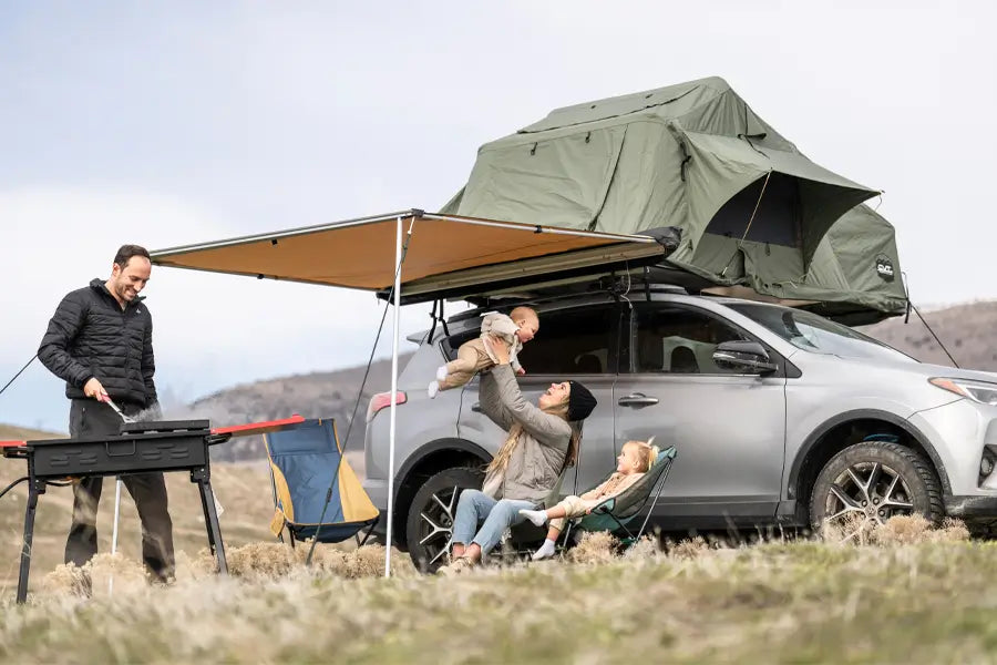 A family relaxing under an awning accessory attached to a rooftop tent above an SUV, while a man cooks on a grill.