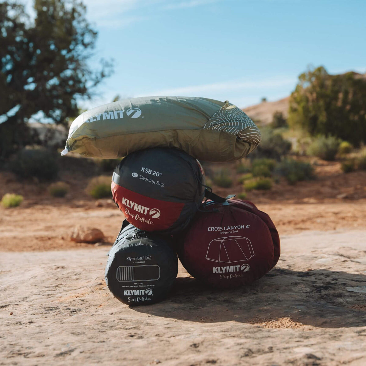 Four packed Klymit sleeping bags stacked outdoors on a rock amid desert vegetation and blue sky, accompanied by a Klymit Drift™ Summit Camp Pillow—water resistant and perfect for your next adventure.