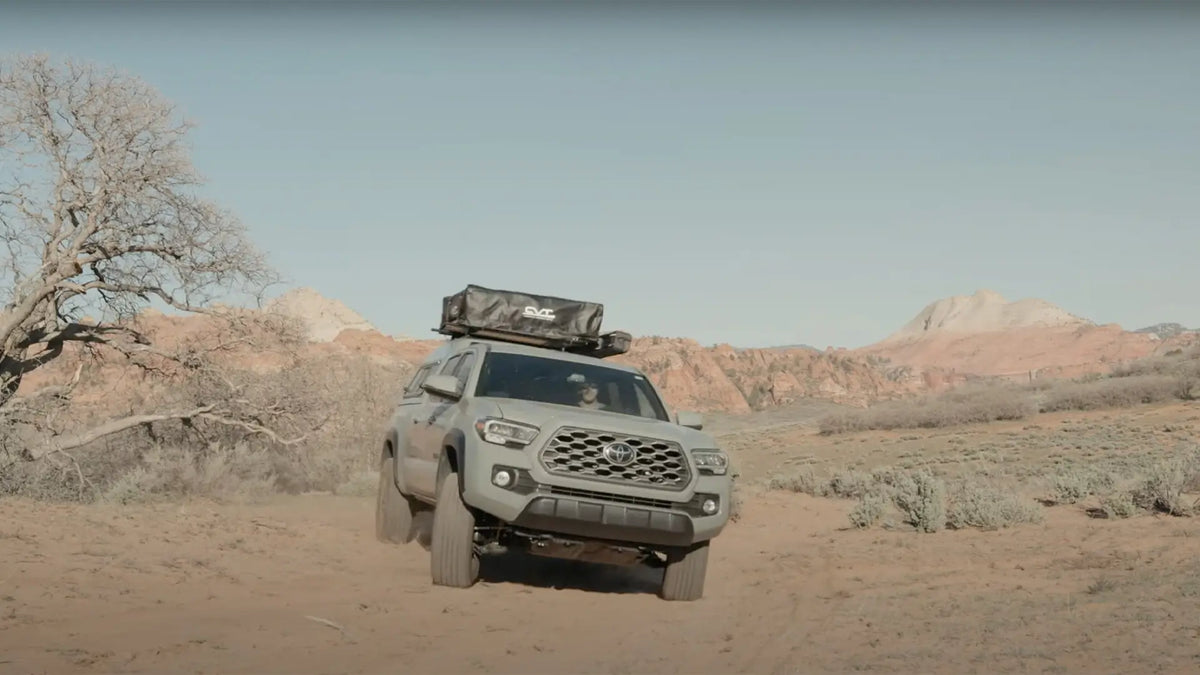 A gray off-road vehicle with a roof tent drives on a dirt path through a desert landscape, surrounded by dry bushes, leafless trees, and distant rocky hills under a clear sky.