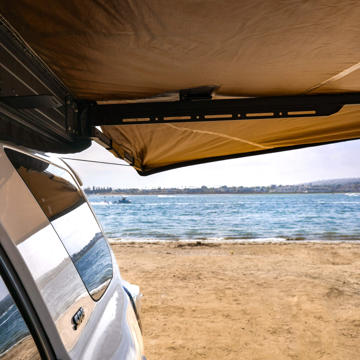 A vehicle equipped with a CVT 180 Degree Awning is parked on a sandy beach, overlooking a calm body of water and distant hills under a partly cloudy sky.
