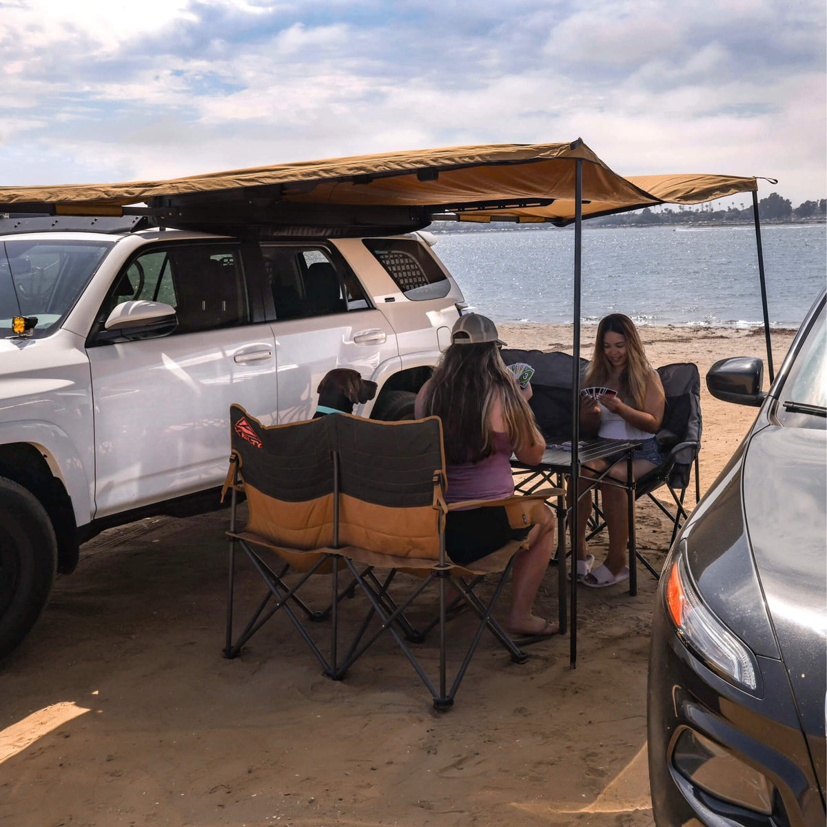 Three people and a dog relax under a CVT 180 Degree Awning mounted to a white SUV, playing cards at a table on the sandy beach. Another car is parked nearby, with the ocean and a partly cloudy sky in the background.