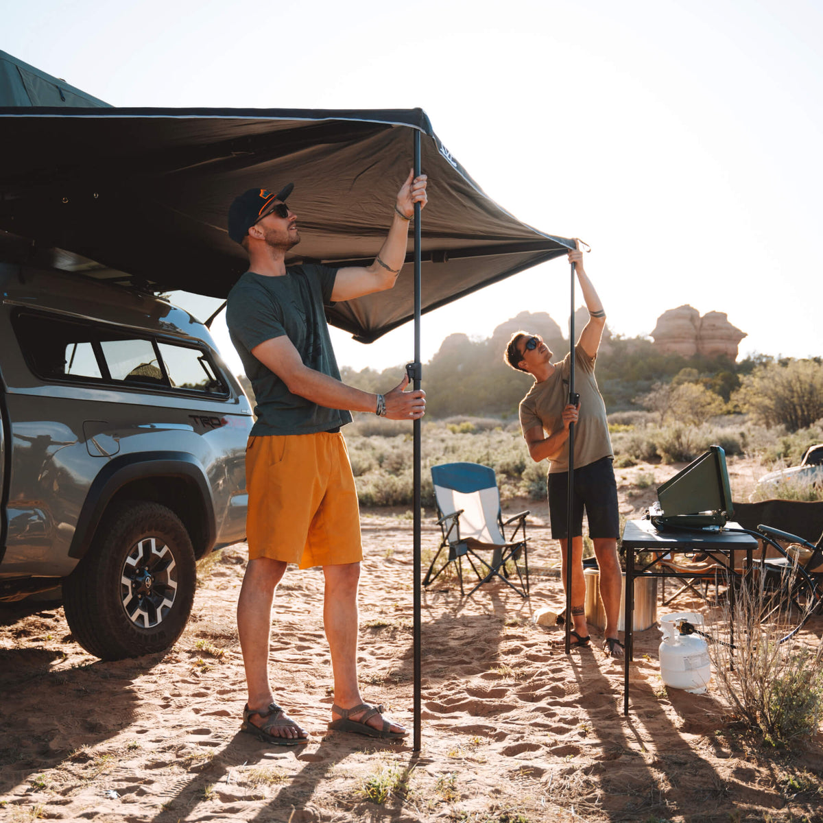 Two people set up a CVT 180 Degree Awning attached to a parked car at a campsite, with chairs, a table, and cooking gear arranged nearby. Sunlight illuminates the desert landscape and distant rock formations.