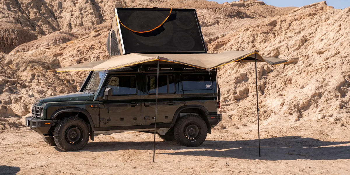 A black off-road SUV is parked on sandy terrain with a rooftop tent open and a large tan awning extended, providing shade. Rocky hills are visible in the background under a clear sky.