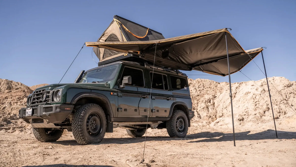 A green off-road SUV is parked in a desert landscape with a rooftop tent and large awning set up, providing shade. The sky is clear and blue.