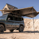 A dark green off-road vehicle equipped with a rooftop tent and the CVT 180 Degree Awning, which offers instant shade, is parked on sandy terrain beneath a clear blue sky.
