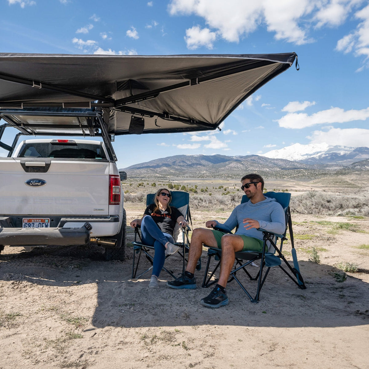 Two people relax in camping chairs under the CVT 270 Degree Awning in a sunny, open landscape with mountains in the distance. They appear to be talking and enjoying the shade, benefiting from reliable four-season weather protection provided by this premium awning from CVT.