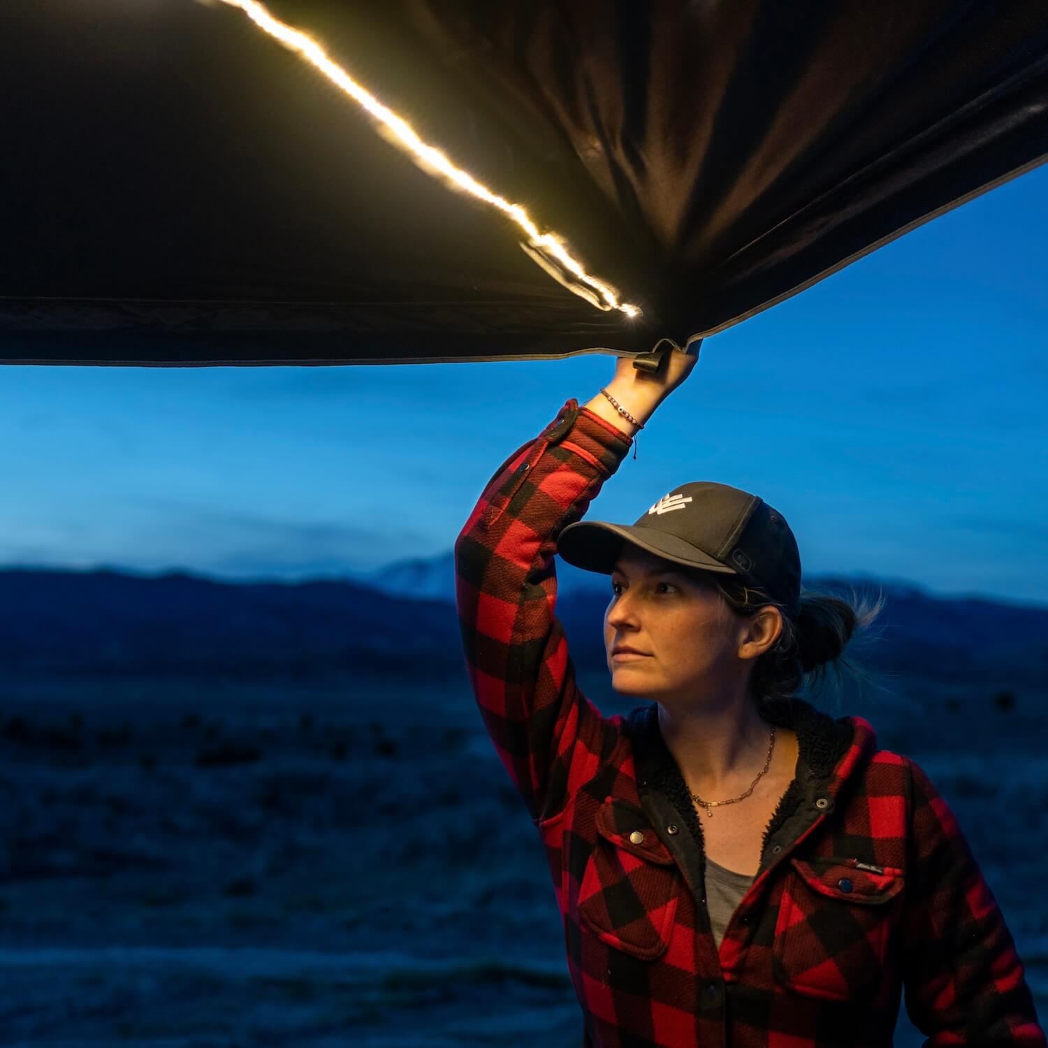 A woman in a red flannel shirt and cap lifts a tent flap with LED lights above her at dusk, with mountains and a darkening sky in the background.