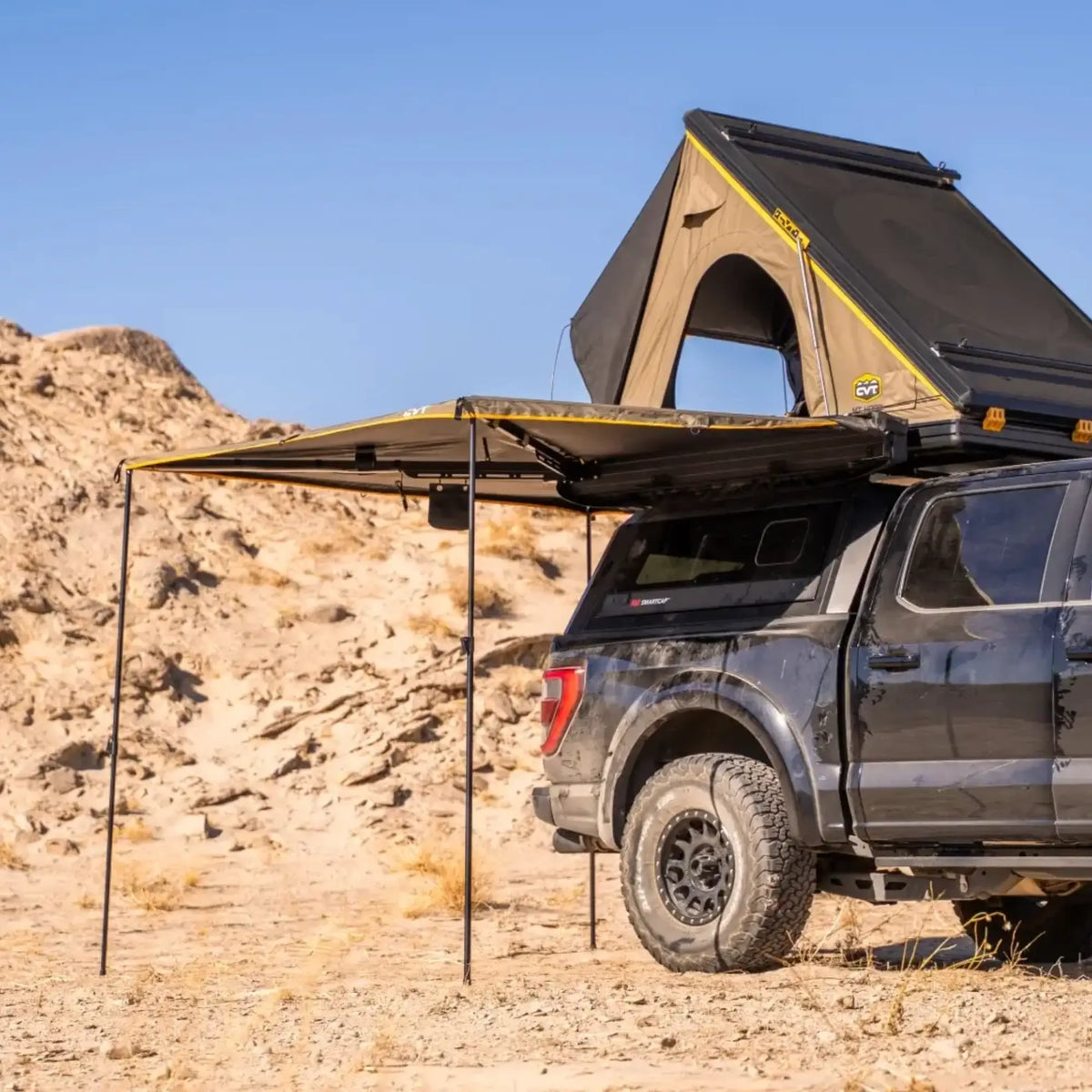 A black truck equipped with a CVT 270 Degree Awning in a batwing design and a rooftop tent is parked on sandy terrain near rocky hills under a clear blue sky.