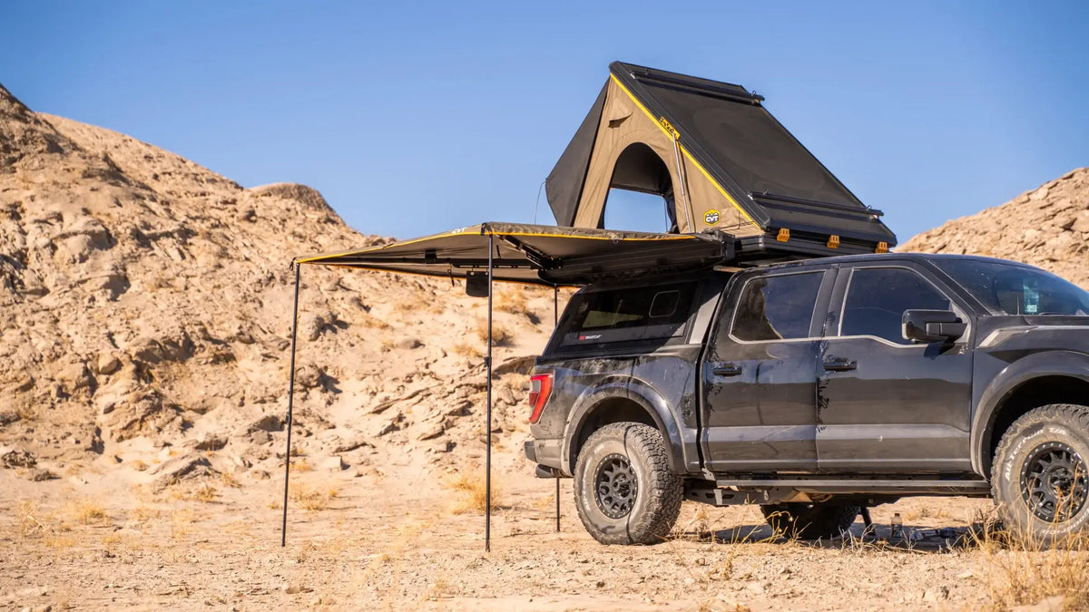 A black pickup truck with a rooftop tent and side awning is parked in a rocky desert landscape under a clear blue sky.