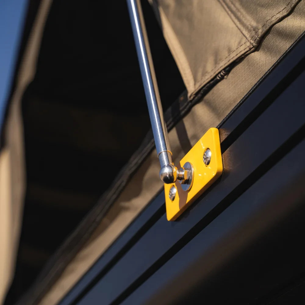 Close-up of a metal tent support pole attached to a yellow mounting bracket on a dark surface, holding up a tan fabric canopy—highlighting the strength and reliability of the CVT Mt. Hood Single Gas Strut.